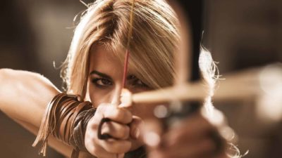 a female archer looking rustic and slightly dishevelled is in extreme close up as she draws back her bow and narrows her eye to aim for a target .