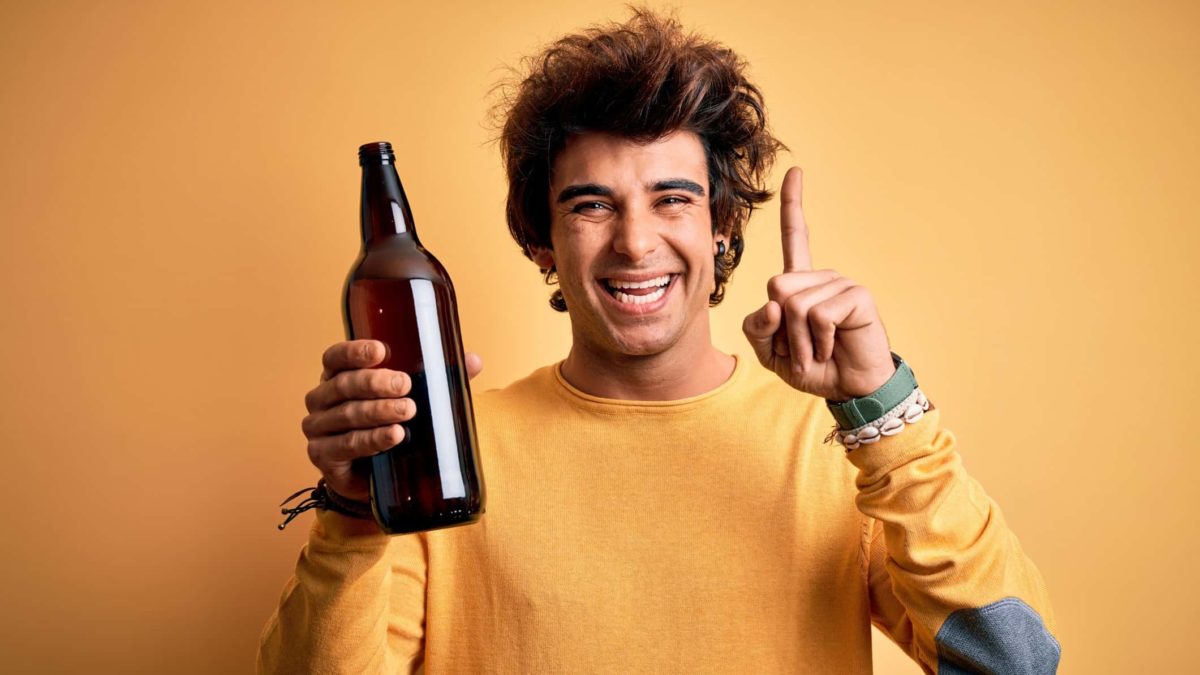 a young man with a wide smile holds a glass bottle in one hand and holds his pointer finger up with the other hand as if indicating a successful outcome.