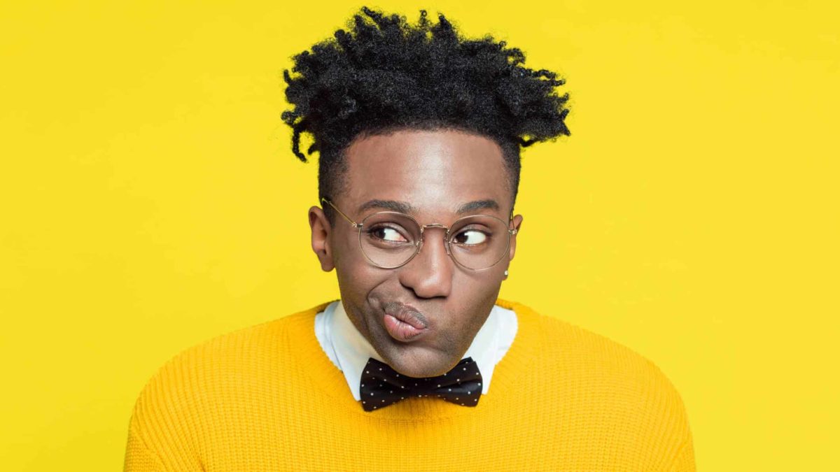 A young man wearing a bright yellow jumper and glasses purses his lips together and moves them to the side of his face as he wonders about something.