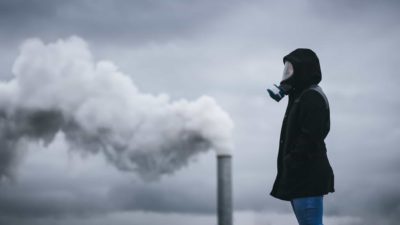 a person stands wearing a full old fashioned gas mask in the foreground of a coal fired smoke stack with smoke billowing into a grey sky and the person standing wearing a coat with hands in pockets.