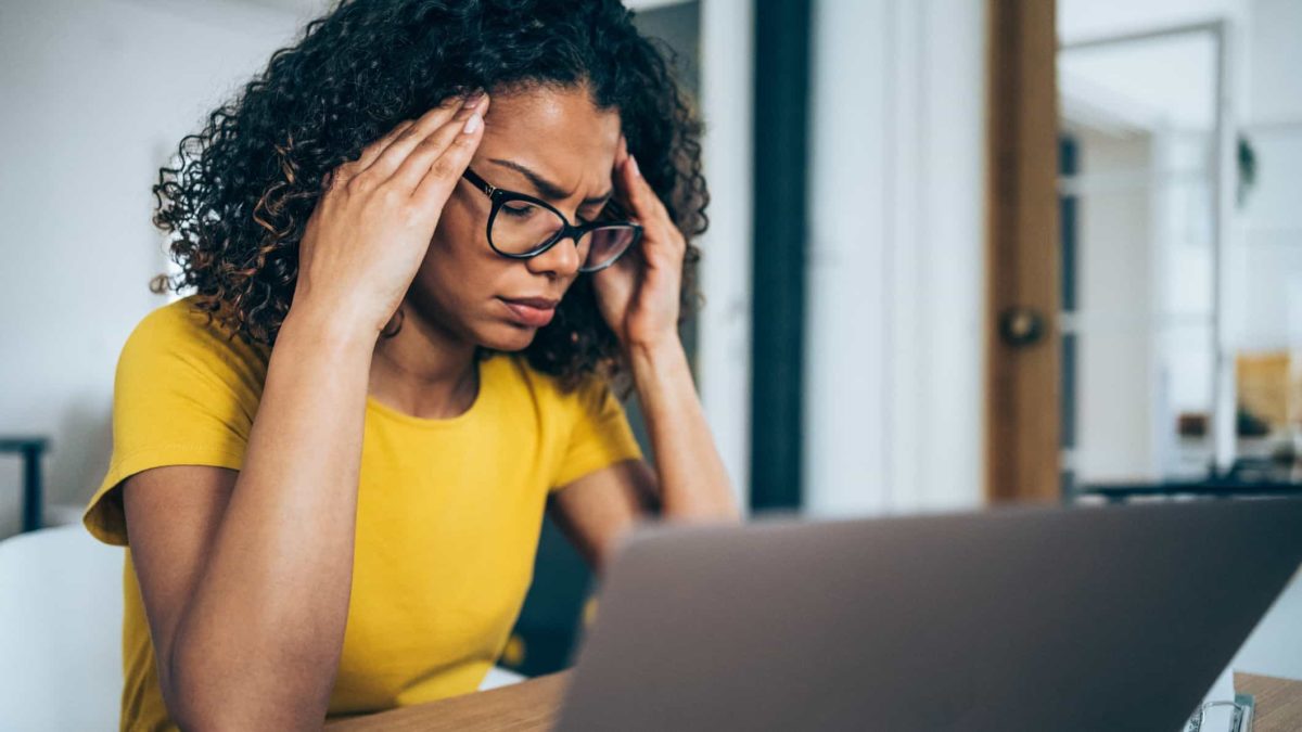 a woman holds her hands to her temples as she sits in front of a computer screen with a concerned look on her face.