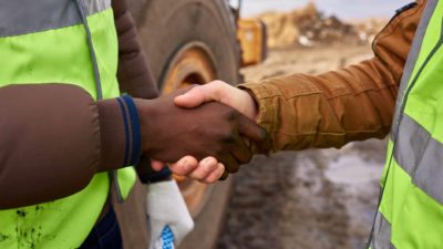 two hands shake in close up at the side of a mine. One party is wearing high visibility gear and there is earth and heavy moving equipment in the background.