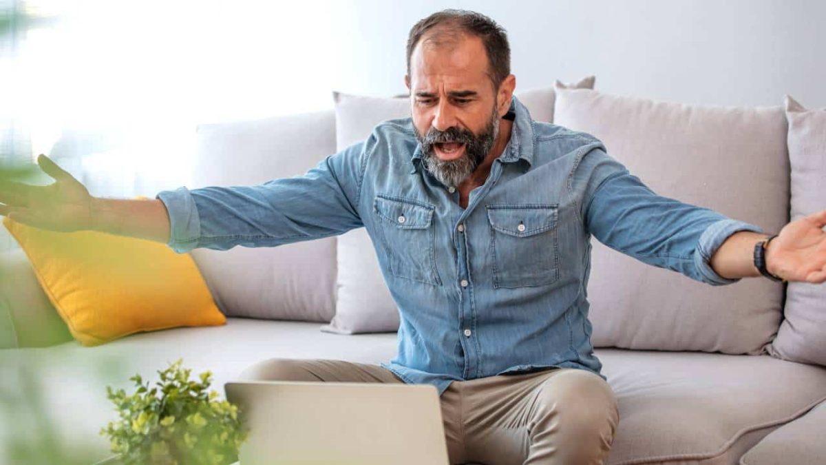 A man sits on a couch with his arms out feeling exasperated while looking at the Costa share price going down on his laptop today