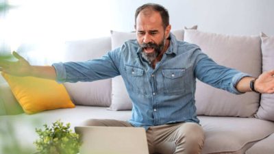 A man sits on a couch with his arms out feeling exasperated while looking at the Costa share price going down on his laptop today