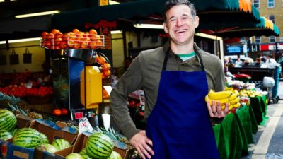 a market stall operator smiles broadly while holding a bunch of bananas with an array of fresh and colourful fruit in the background.