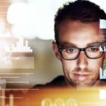 a man wearing spectacles has a satisfied look on his face as he appears within a graphic image of graphs, computer code and technology related symbols while he concentrates on a computer screen
