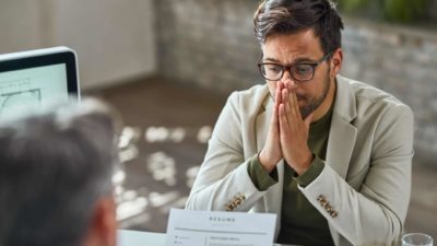 a man in a suit jacked sits uncomfortably with his hands clasped before his face in a job interview situation while sitting across from an interviewer