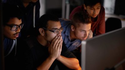A group of people gather around a computer screen in rapt attention, one man holds his hands to cover his mouth as if in nervous anticipation of what news may come.