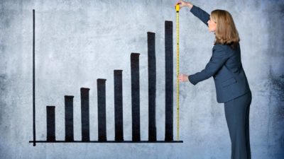 a woman in a business suit uses an old fashioned tape measure holding it up against the tallest bar in a bar graph on a wall.