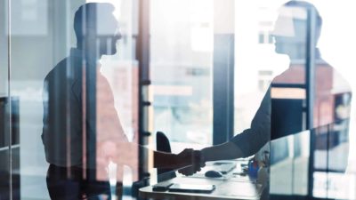 Two businessmen shake hands behind a window.