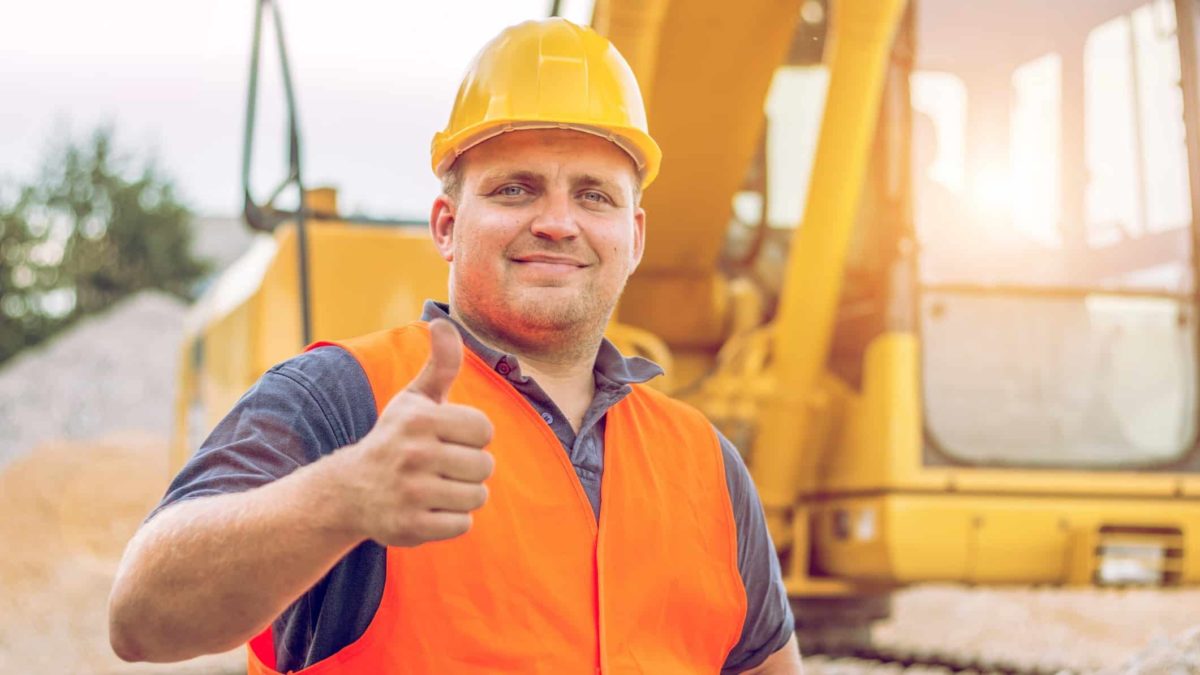 A man in a hard hat and high visibility vest holds his thumb up in a gesture of confidence with heavy moving equipment in the background as on a mine site as the Chalice Mining share price rises today