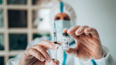 a medical person in full protective gear with mask and gloves holds up a needle in one hand and a small bottle of vaccine in the other in a medical setting.