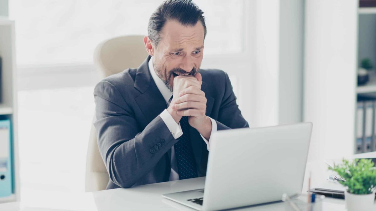 a man weraing a suit sits nervously at his laptop computer biting into his clenched hand with nerves, and perhaps fear.