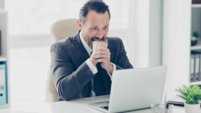 a man weraing a suit sits nervously at his laptop computer biting into his clenched hand with nerves, and perhaps fear.