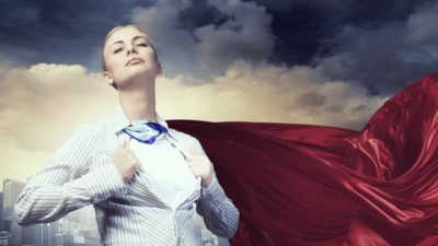 A woman in a suit pulls apart shirt and wears cape while looking strong in front of city skyline.
