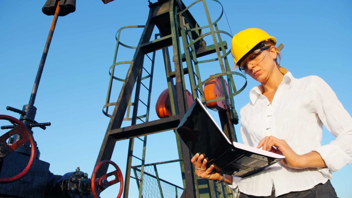 an oil refinery worker checks her laptop computer in front of a backdrop of oil refinery infrastructure. The woman has a serious look on her face.