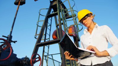 an oil refinery worker checks her laptop computer in front of a backdrop of oil refinery infrastructure. The woman has a serious look on her face.