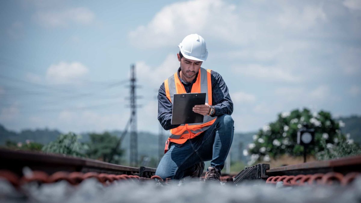 a railway worker squats down in between tracks to note something on his documentation. He is waring a hard hat and high visibility vest and there is signalling equipment in the background.