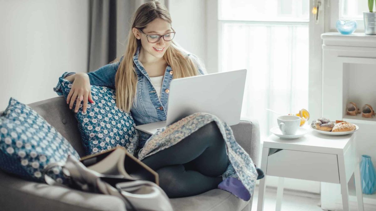 A young woman sits on a sofa in a stylish home with her laptop computer balanced on her knee and smiles with a satisfied look on her face at what she's seeing on the screen.