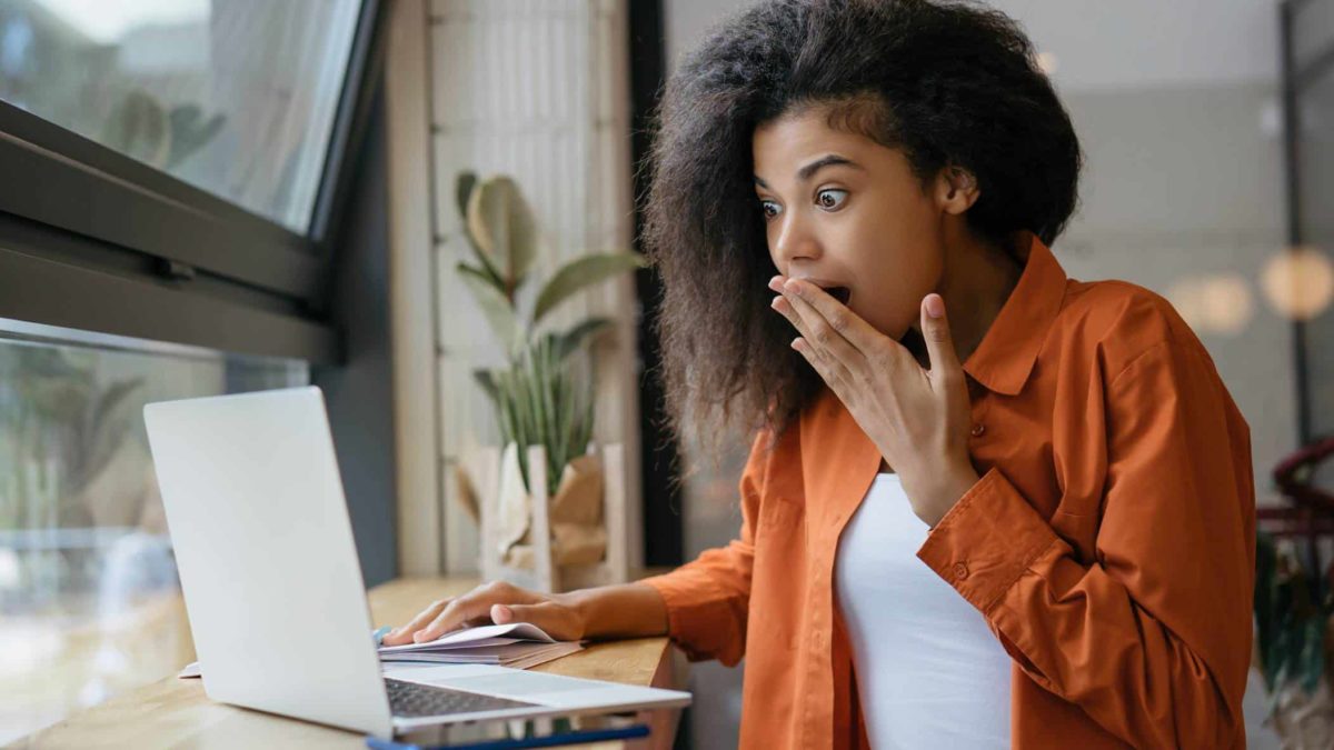 A young woman holds her hand to her mouth in surprise as she reads something on her laptop.