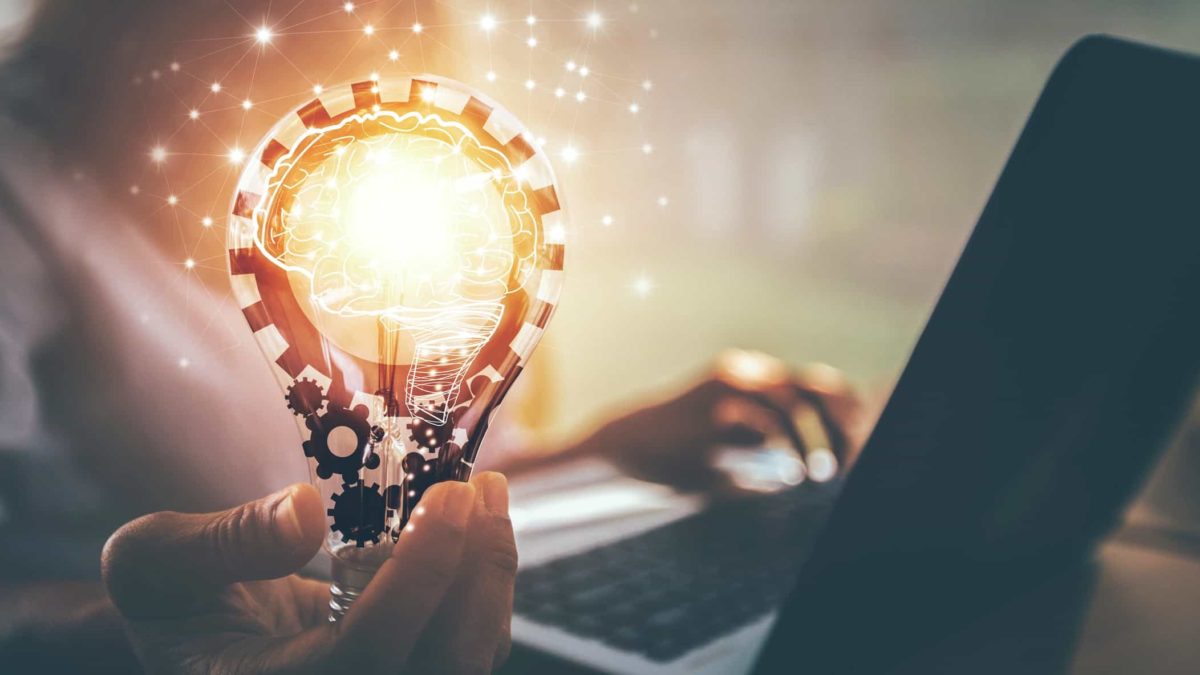 A person working on a computer holds a lightbulb that is connected to the network and shining brightly.