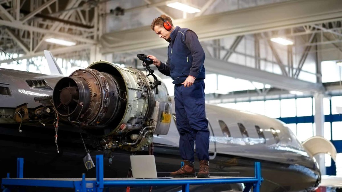 an aircraft maintenance technician stands atop a platform inspecting the jets of an aircraft within a hangar.