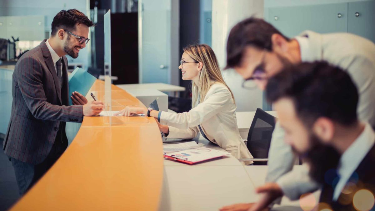 a group of four people in a bank setting with one woman serving a customer and the other two male bank workers grouped together over a document.