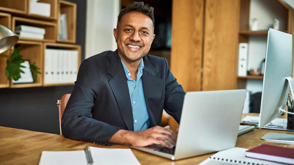 a man in a business suit sits at his laptop computer at his desk and smiles broadly in an office setting, giving an air of optimism and confidence.