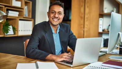 a man in a business suit sits at his laptop computer at his desk and smiles broadly in an office setting, giving an air of optimism and confidence.