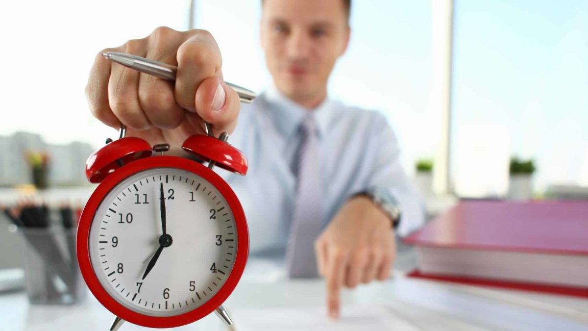 A man points at a paper as he holds an alarm clock.