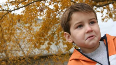 a young boy with a zip-up jacked stands in front of a large tree limb full of autumn-coloured leaves.