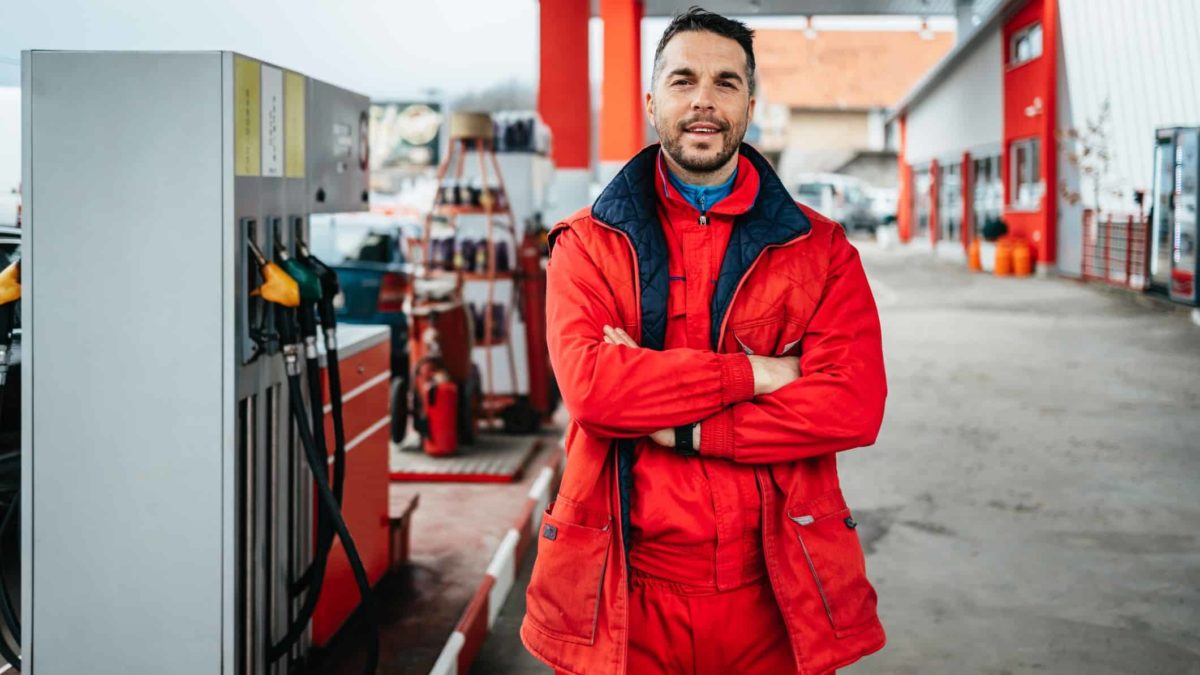 a service station attendant crosses his arms and smiles towards the camera with a backdrop of petrol bowsers and a drive-through facility.