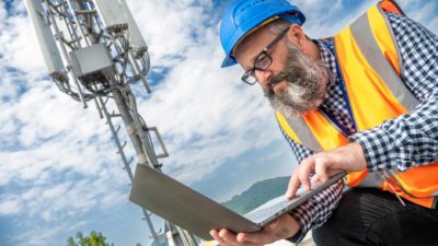 a telecommunications technician checks his laptop computer while wearing a hard hat and a high visibility vest with a large mobile phone installation tower in the background of the picture.