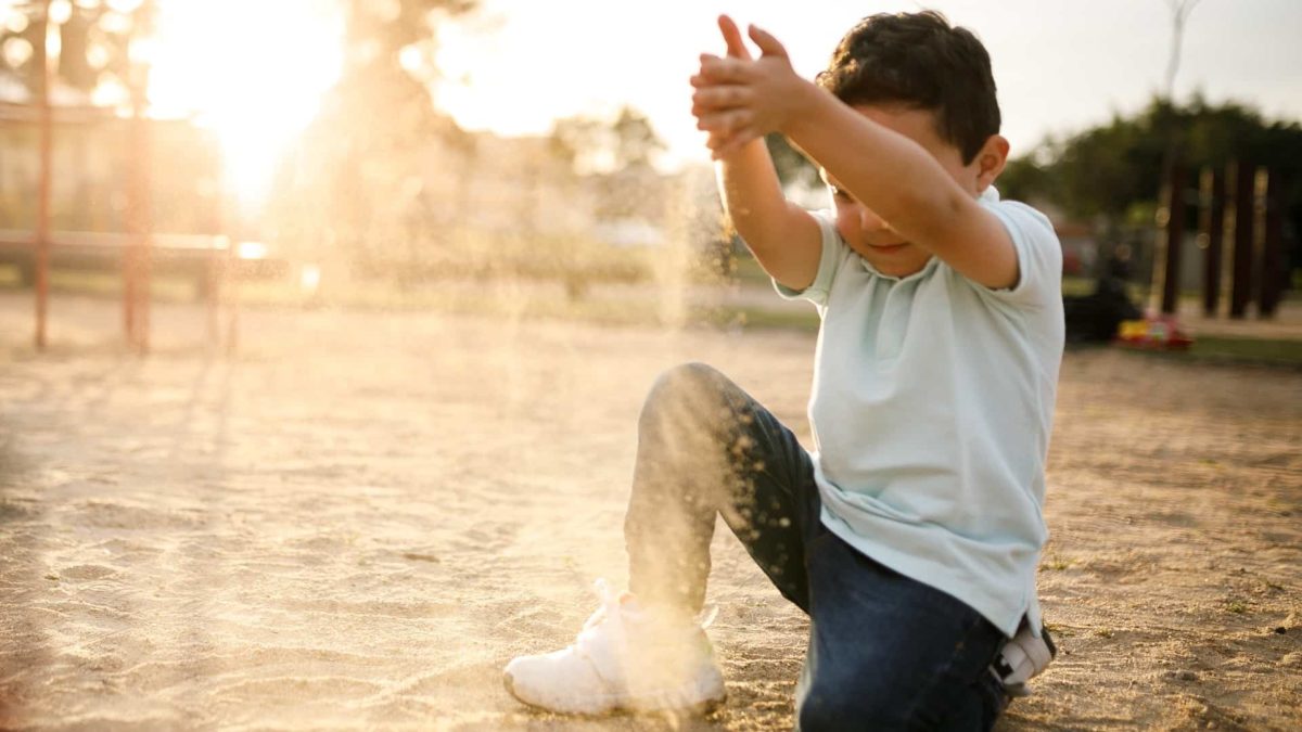 A small child in a sandpit holds a handful of sand above his head and lets it trickle through his fingers.