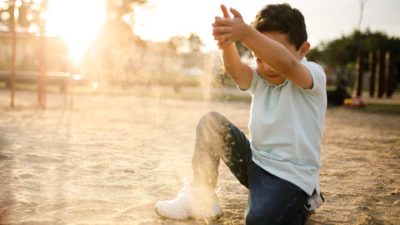 A small child in a sandpit holds a handful of sand above his head and lets it trickle through his fingers.