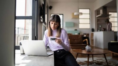 a woman in a stylish living space stands leaning into her laptop computer with credit card in hand as though she is online shopping.