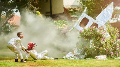 Two children dressed as space travellers in white suits look on at the smoking wreckage of their tin foil covered carboard rocket in their backyard with one child pulling the other away from the crash site.