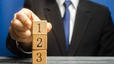 A man in a business suit and tie places three wooden blocks with the numbers 1, 2 and 3 on them on top of each other on a table. representing the most traded ASX 200 shares by volume today