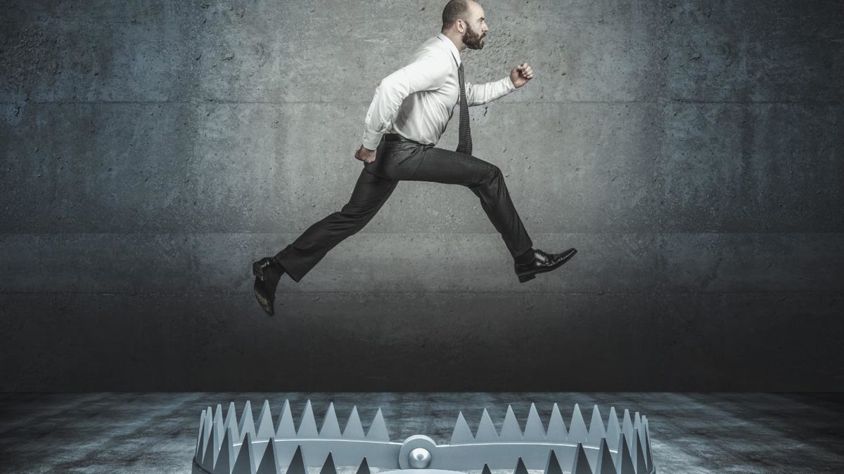 a man in a business shirt and tie takes a wide leap over a large steel trap with jagged teeth that is place directly underneath him.