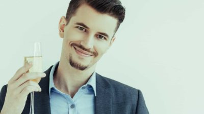 a young man wearing an open necked shirt and a stylish coat raises a glass of champagne as he smiles.