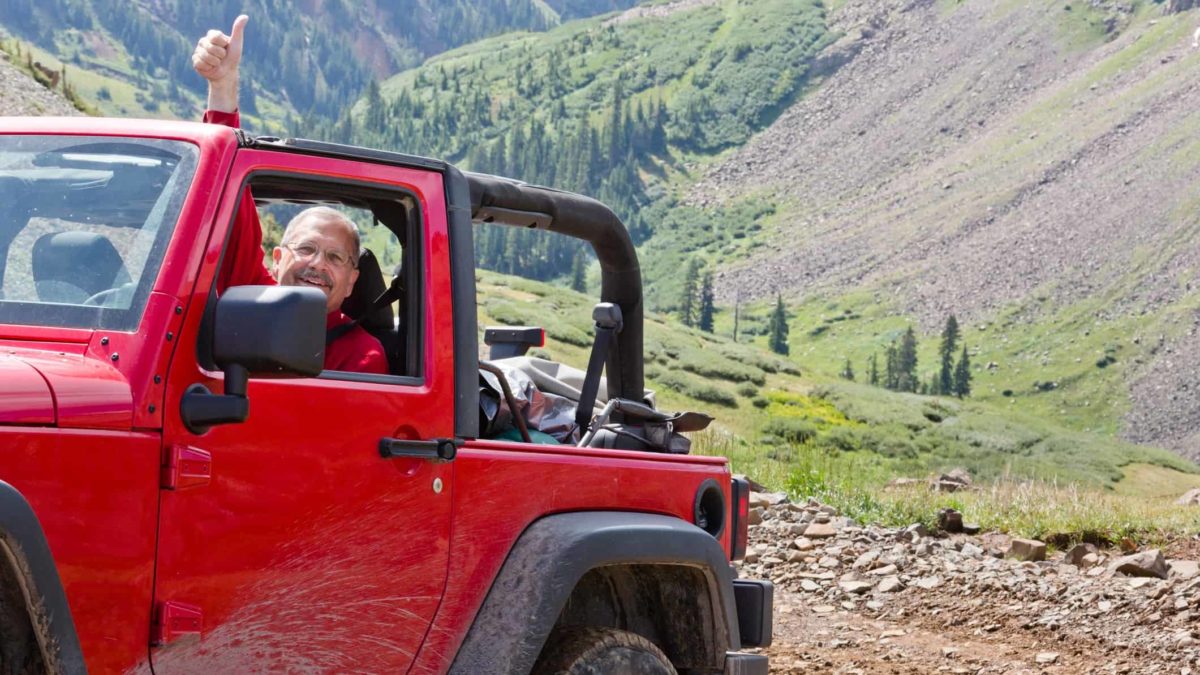 a man in a four wheel drive vehicle lifts an arm and gives a thumbs up in the air as he traverses rugged mountrain style terrain with a green valley and rocky hills in the background.