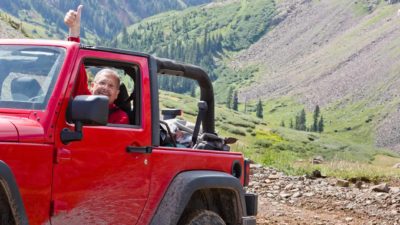 a man in a four wheel drive vehicle lifts an arm and gives a thumbs up in the air as he traverses rugged mountrain style terrain with a green valley and rocky hills in the background.