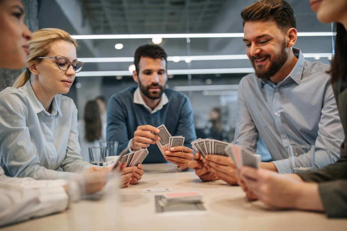 A group of people sit around a table playing cards in a work office style setting.