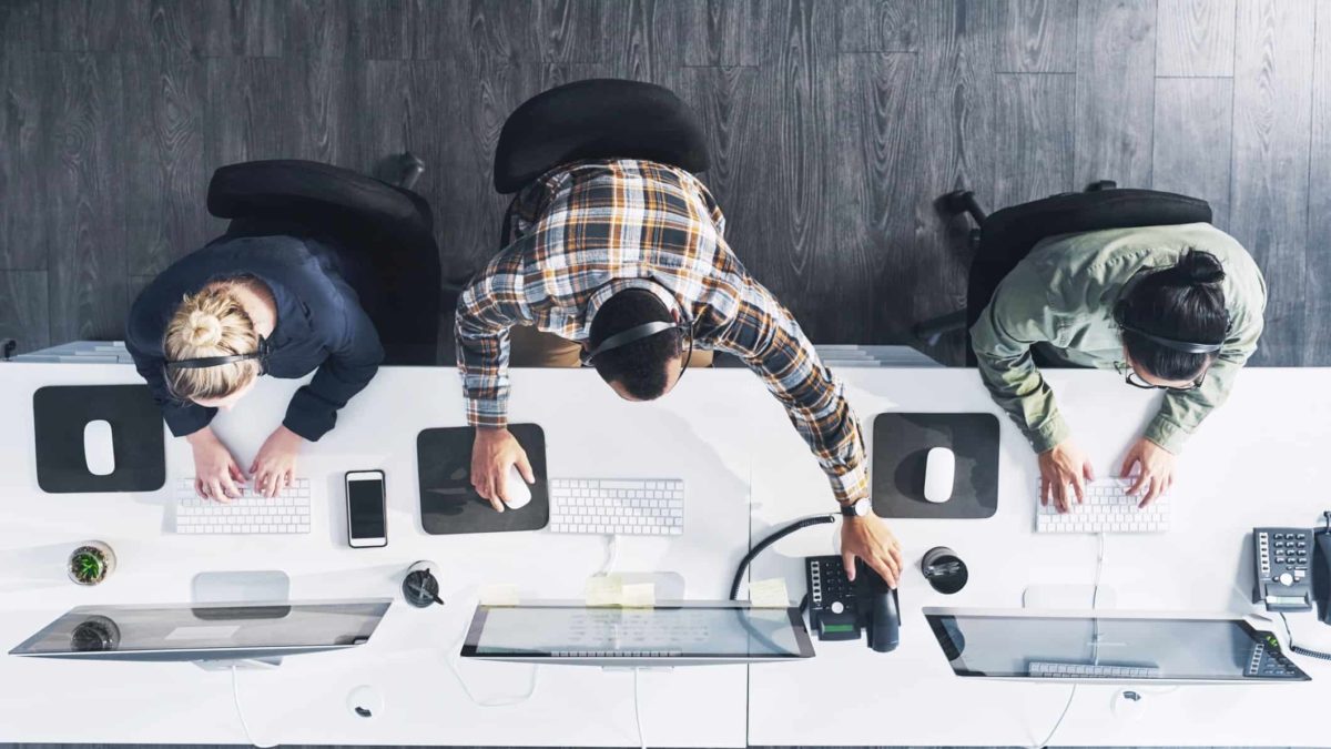 Looking down on a workstation with three people working on their tech devices.