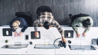 Looking down on a workstation with three people working on their tech devices.