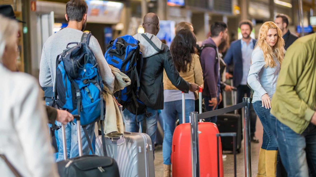 a crowd of people at an airport stand, some in queues, others looking around, while all drag their bags on wheels beside them.