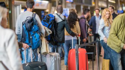 a crowd of people at an airport stand, some in queues, others looking around, while all drag their bags on wheels beside them.