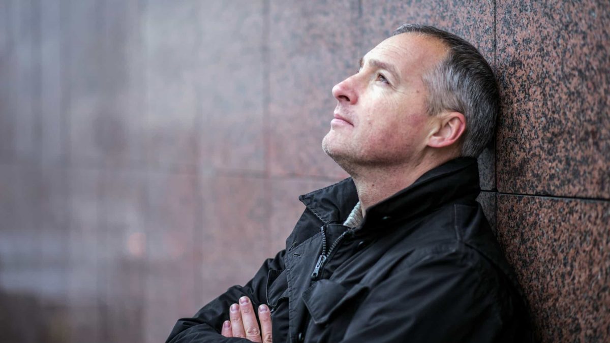 A concerned man leans against a brick wall looking up at the sky
