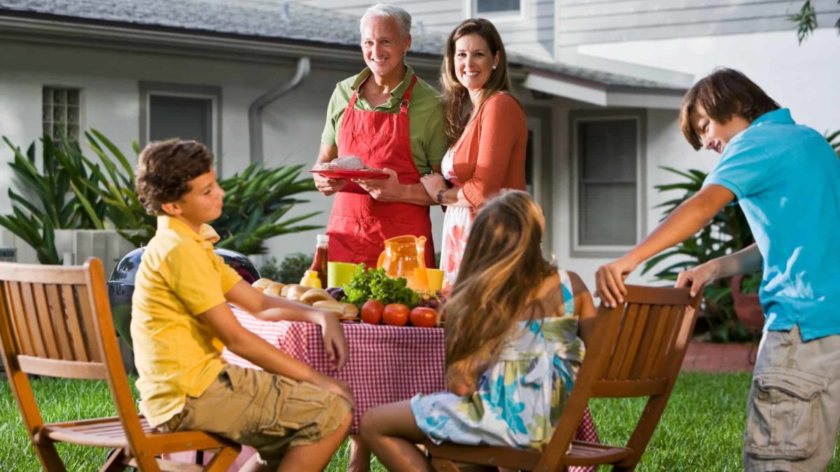 a family has an outdoor table set up, laden with food, as they prepare for a meal outside on the grass with their home in the background.