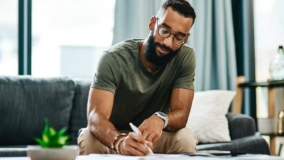 a man sits in a home environment on a sofa while writing in a book with a pen, a plant on the table nearby and curtains open in the background.